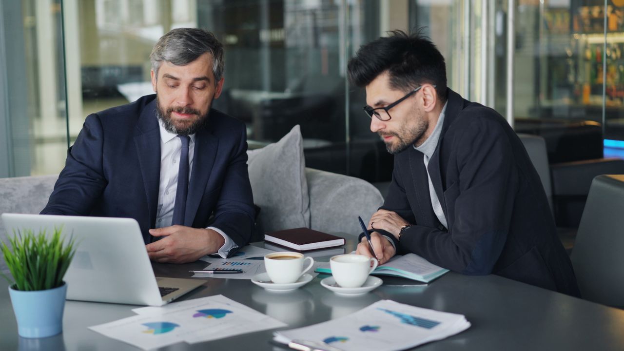 Two businessmen in suits reviewing charts and graphs over coffee in a modern office setting.
